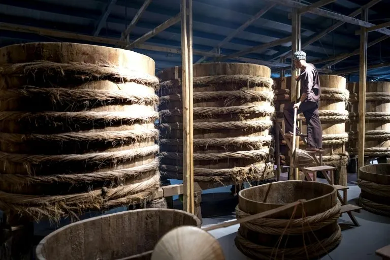 A worker oversees large wooden barrels used for traditional fermentation in an indoor warehouse setting.